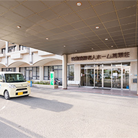 介護施設の玄関・車寄せ。白い送迎車が停まっており、入口ドアと受付窓口が見える。
